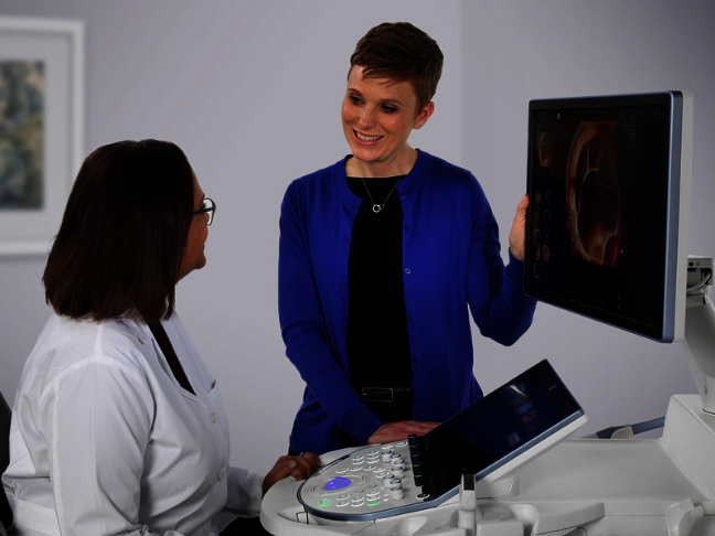 Two women, one of them wearing a white coat are standing next to an ultrasound system and talking to each other