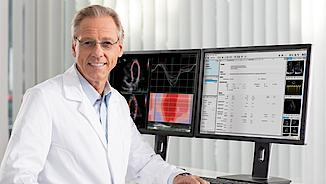 Physician sitting at his desk. Behind him 2 PC monitors with ViewPoint 6 software