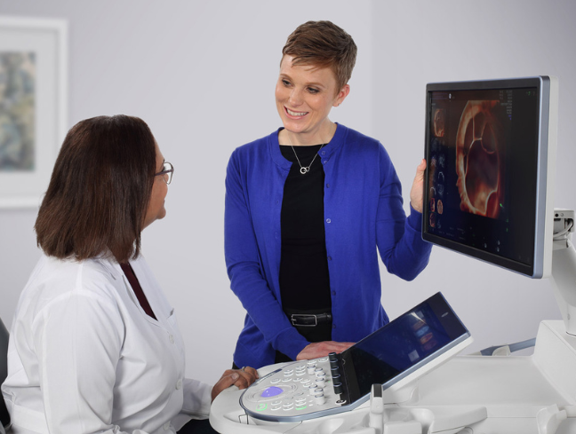 Zwei Frauen, eine davon im Arztkittel, stehen neben einem Ultraschallgerät und unterhalten sich.