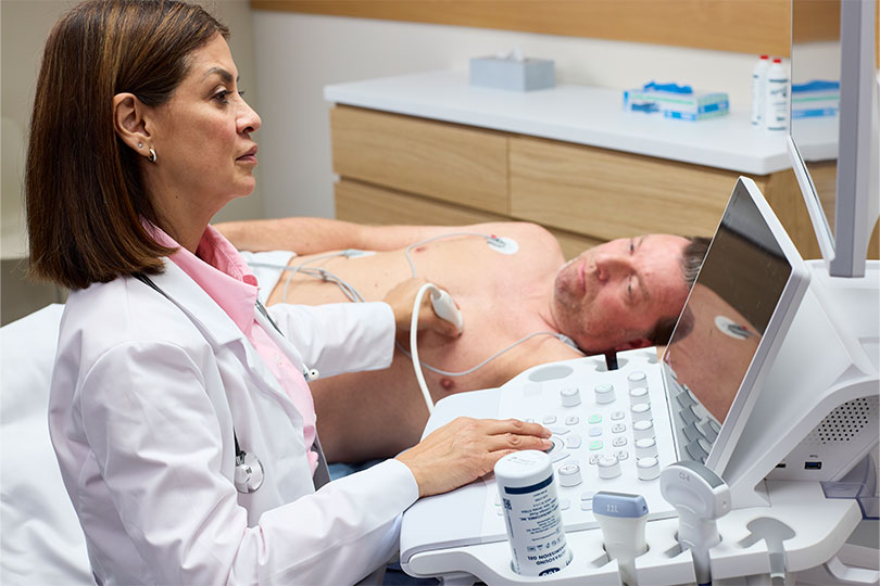 A female doctor operating a GE ultrasound console while performing a cardiac scan on a male patient connected to ECG leads on an examination bed.