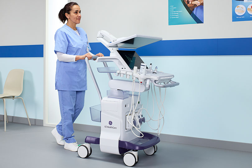 A healthcare professional in blue scrubs pushing a mobile GE Healthcare ultrasound cart down a hospital hallway.