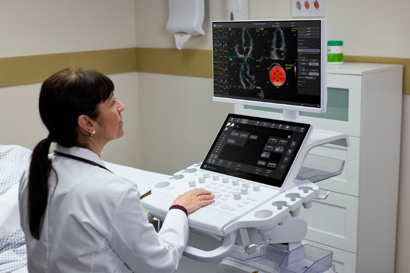 A female doctor in a white coat operating a GE Healthcare ultrasound machine, with the main monitor displaying multi-plane echocardiogram views and a strain bullseye plot.