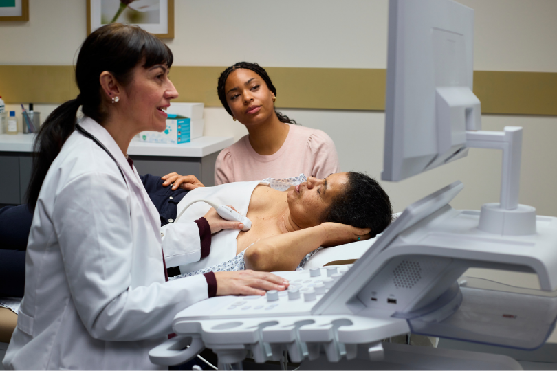 A doctor smiling while performing an ultrasound scan on a patient's chest and abdomen, as an observer sits nearby in the examination room.
